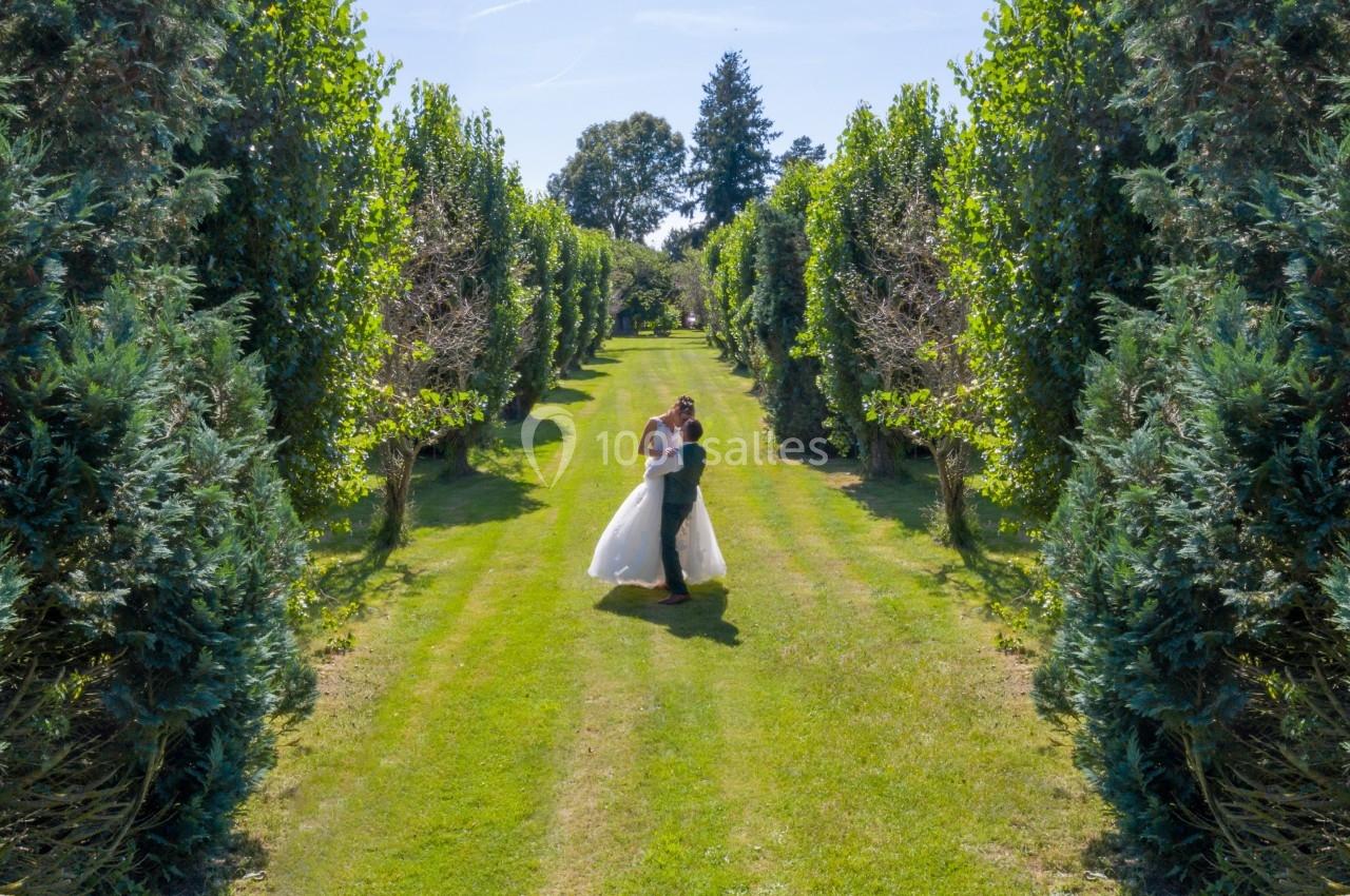 Un couple en tenue de mariage marche sur une allée bordée d'arbres dans un jardin verdoyant.
