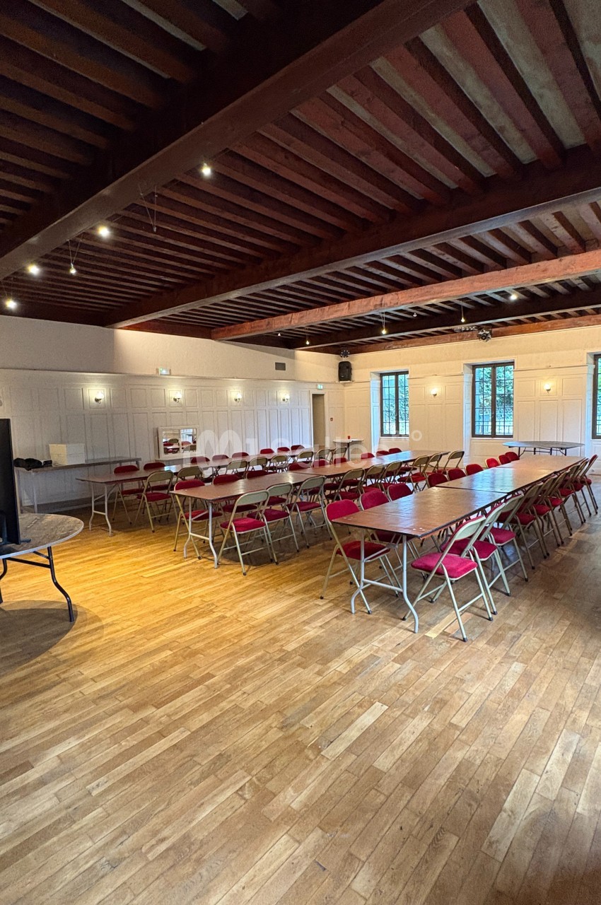 Salle lumineuse avec parquet en bois, tables rectangulaires alignées et chaises rouges, plafond en poutres apparentes.