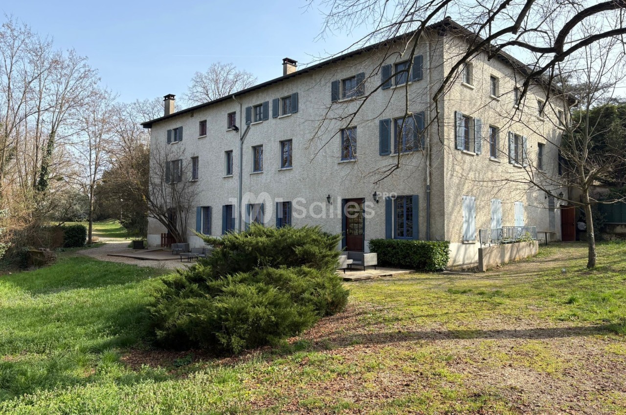 Grande maison blanche à deux étages avec volets bleus, entourée d'arbres et d'un jardin verdoyant.