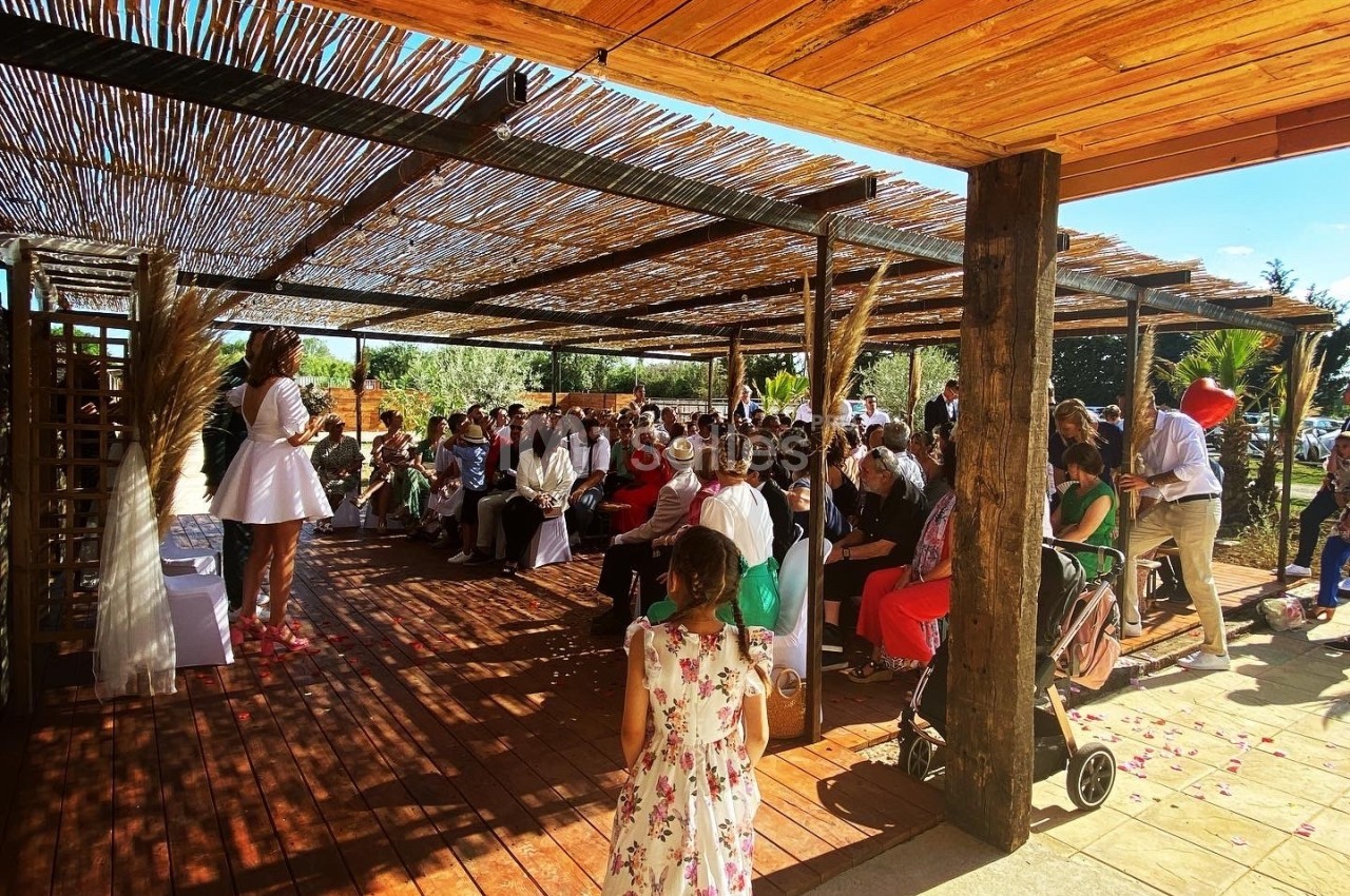 Une femme en robe blanche s'adresse à un public assis sous une pergola en bois lors d'un événement en extérieur.