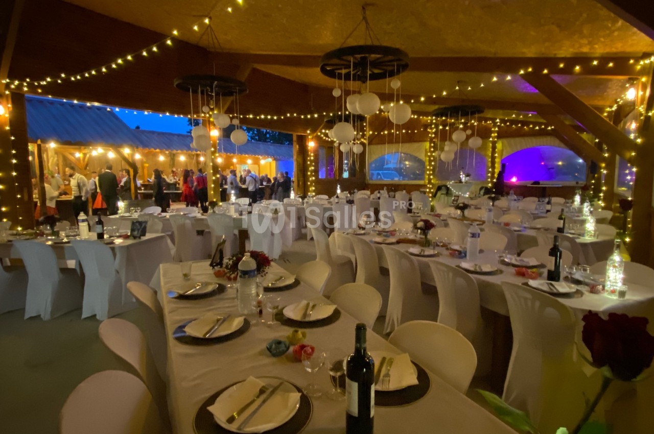 Salle de réception décorée avec des guirlandes lumineuses, tables dressées et invités en arrière-plan.