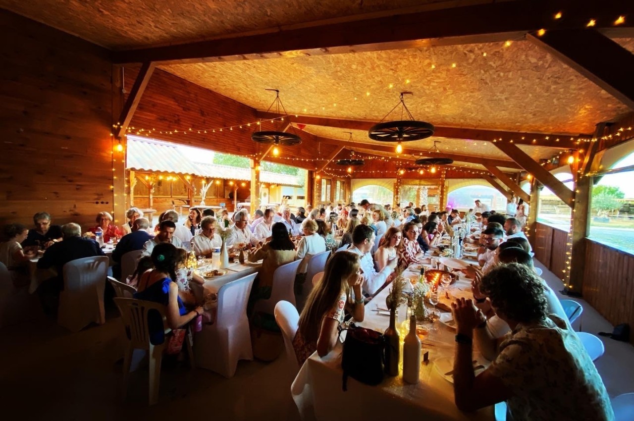 Salle en bois décorée de guirlandes lumineuses, accueillant des convives assis à de longues tables lors d'un événement.