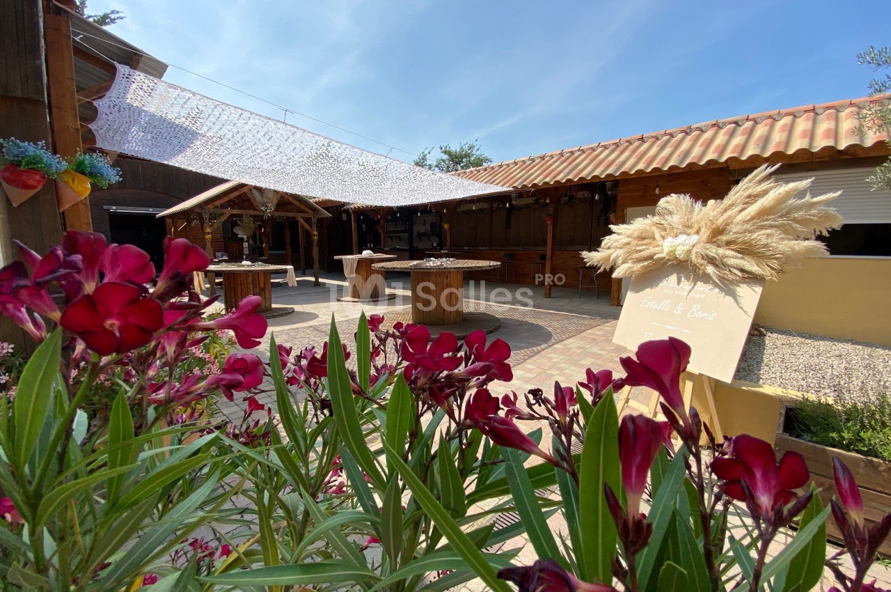 Espace extérieur avec tables en bois, fleurs au premier plan et voile d'ombrage sous un ciel dégagé.