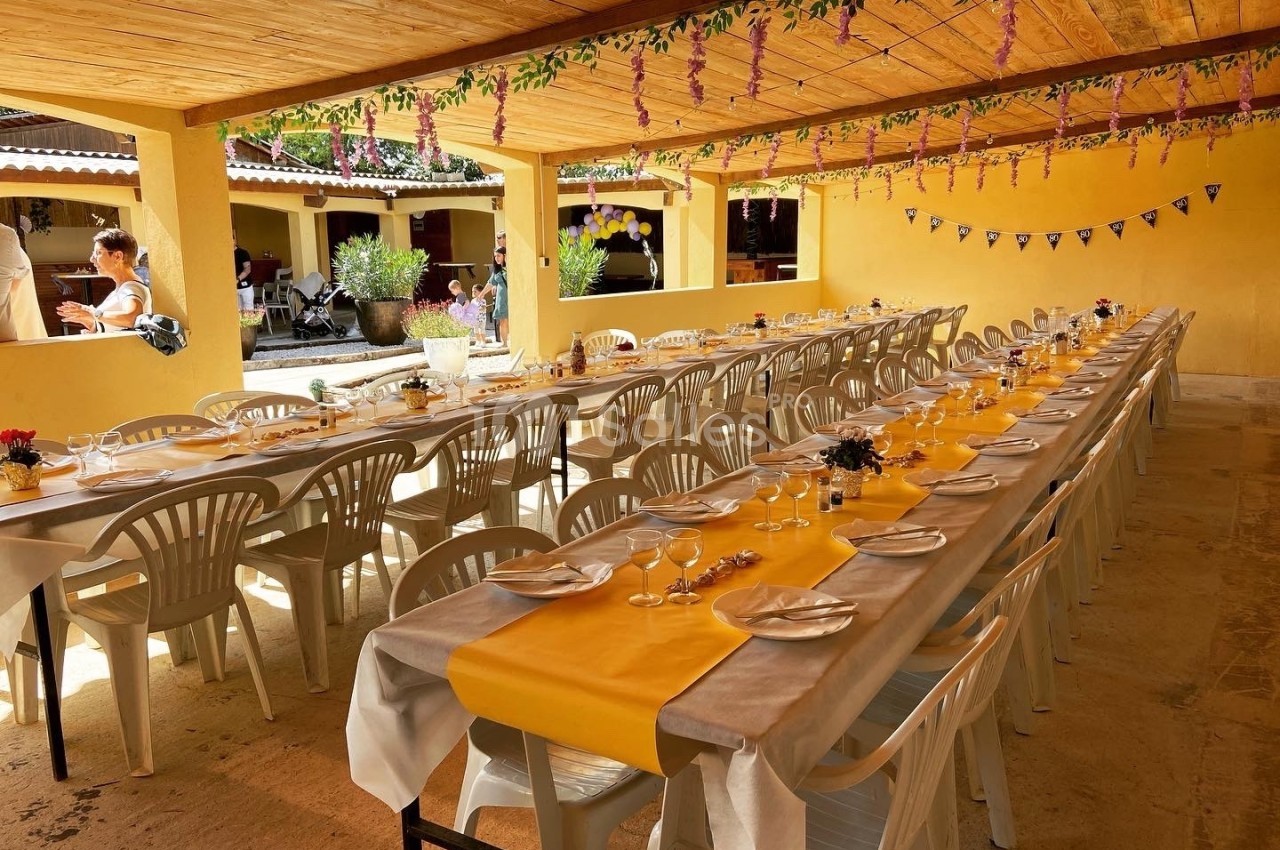 Tables longues dressées avec nappes jaunes et blanches sous un auvent décoré de guirlandes fleuries.