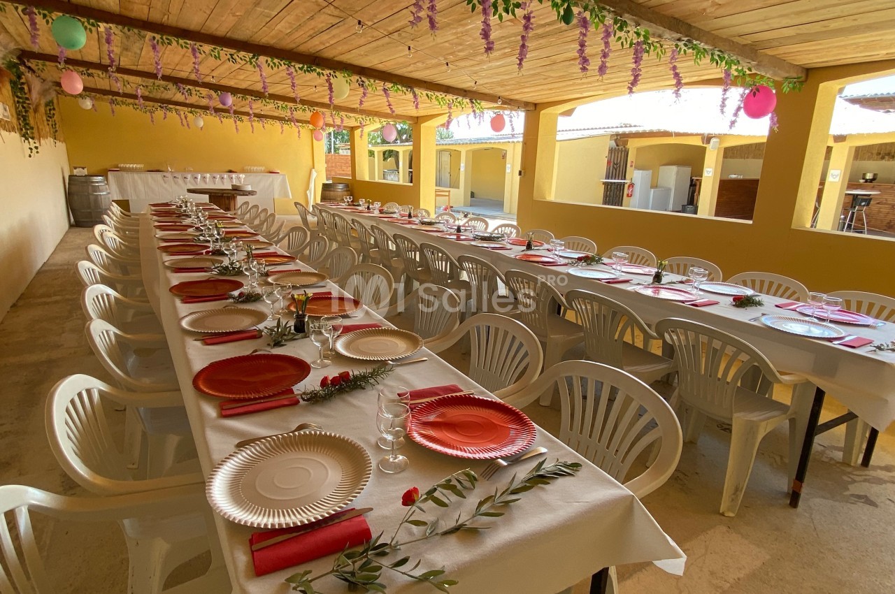 Salle décorée pour un repas festif avec de longues tables alignées, nappes blanches et assiettes colorées.