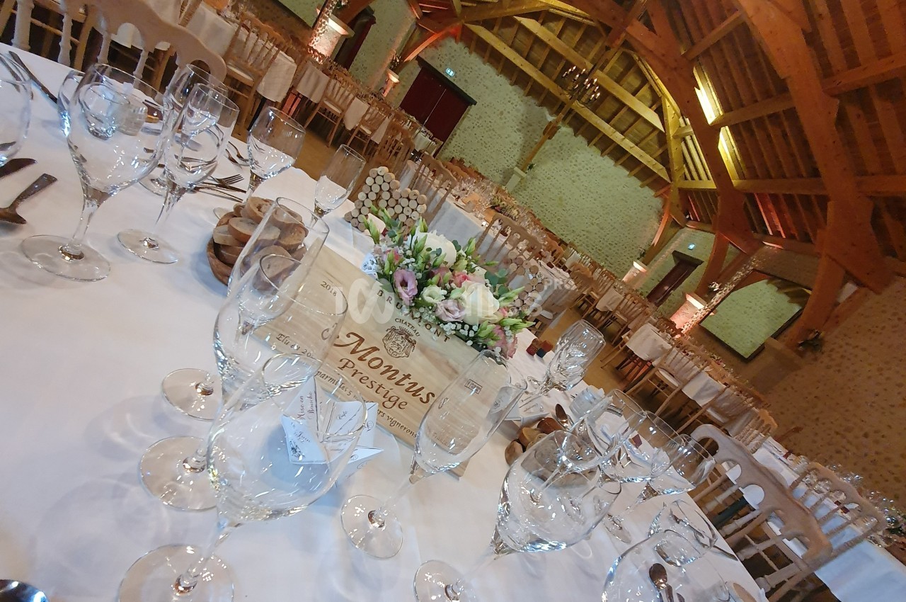 Salle de réception avec tables dressées, nappes blanches, verres, couverts et centre de table floral sous une charpente en…