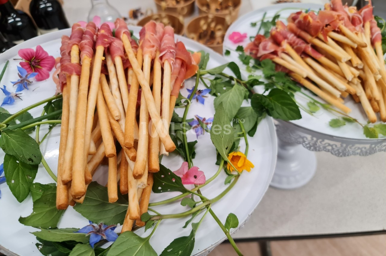 Assortiment de gressins enveloppés de jambon cru, présentés sur des plateaux décorés de feuilles et de fleurs comestibles.