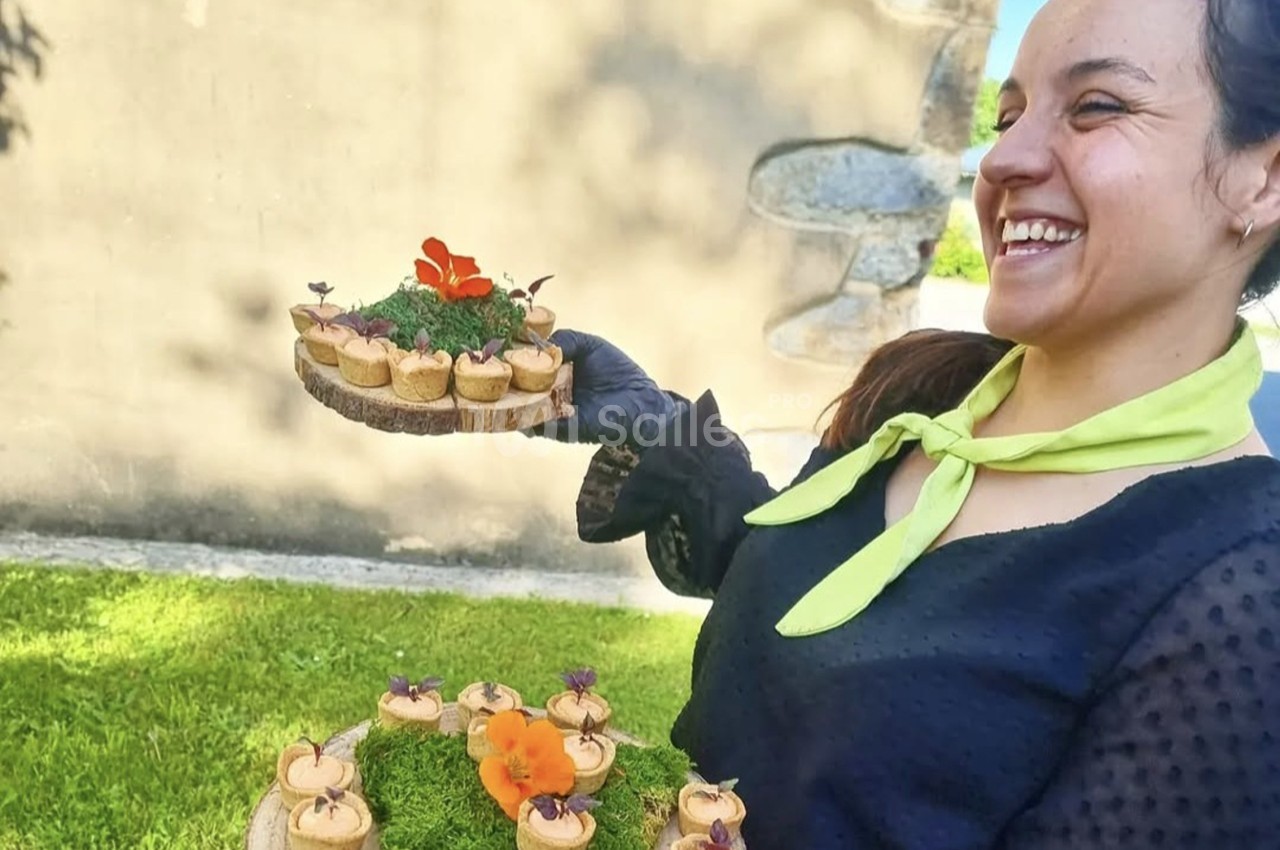 Une femme souriante présente des amuse-bouches décorés de fleurs sur des plateaux en bois, en extérieur.