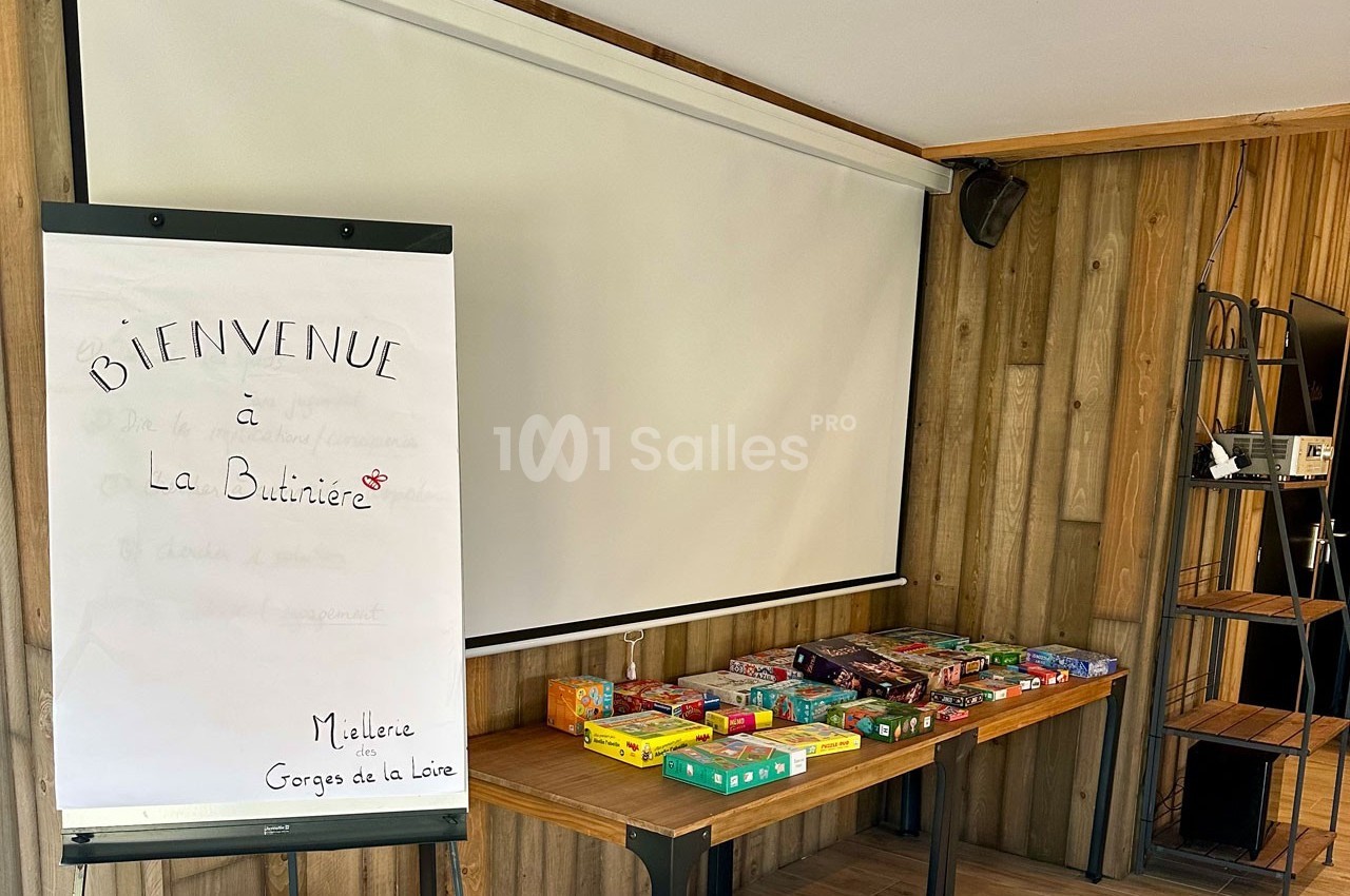 Table avec des jeux de société alignés, tableau de bienvenue écrit à la main et écran de projection dans une salle en bois.