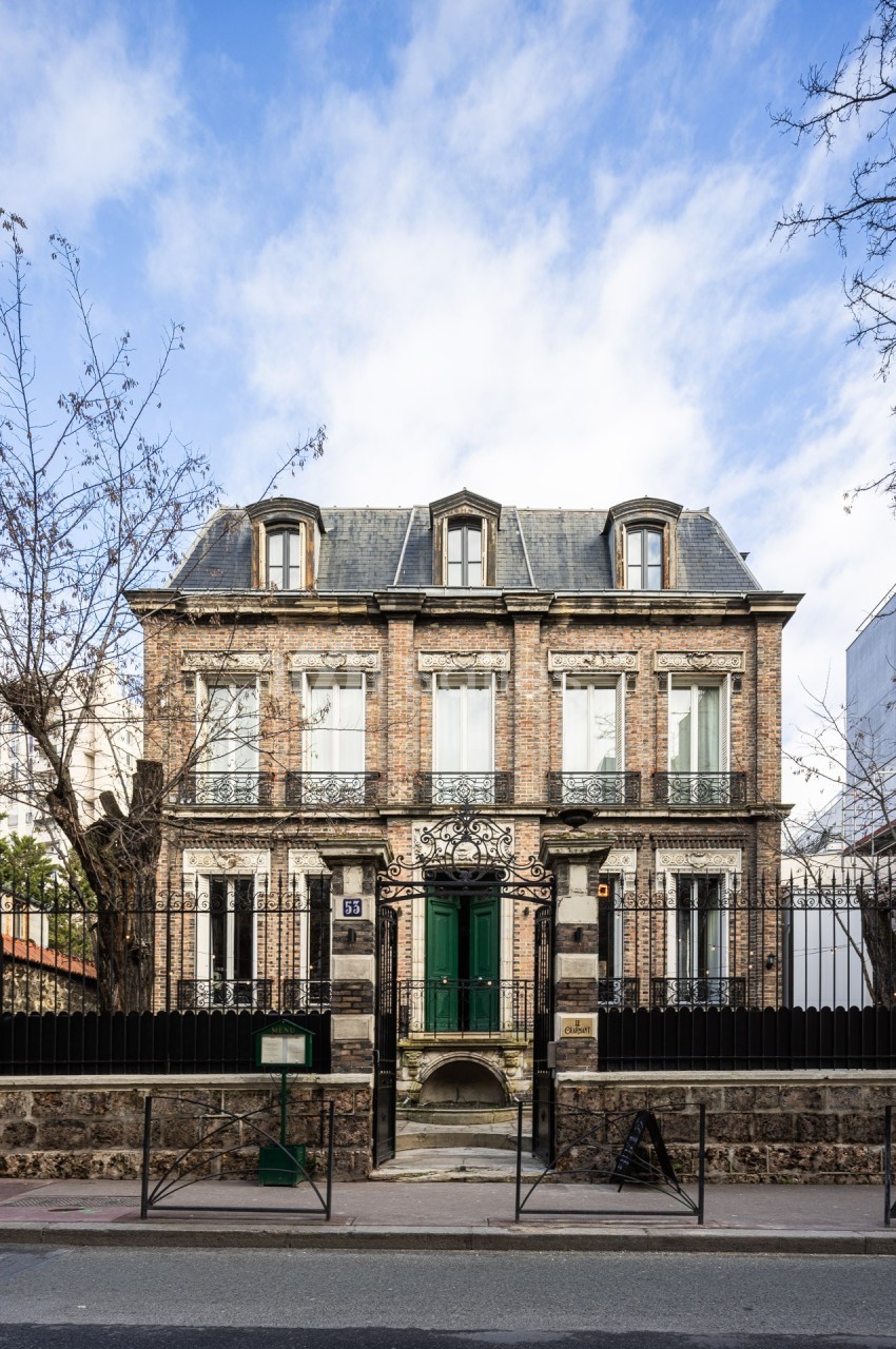 Façade d'une maison en briques à deux étages avec toit mansardé, portail en fer forgé et arbres dénudés devant.