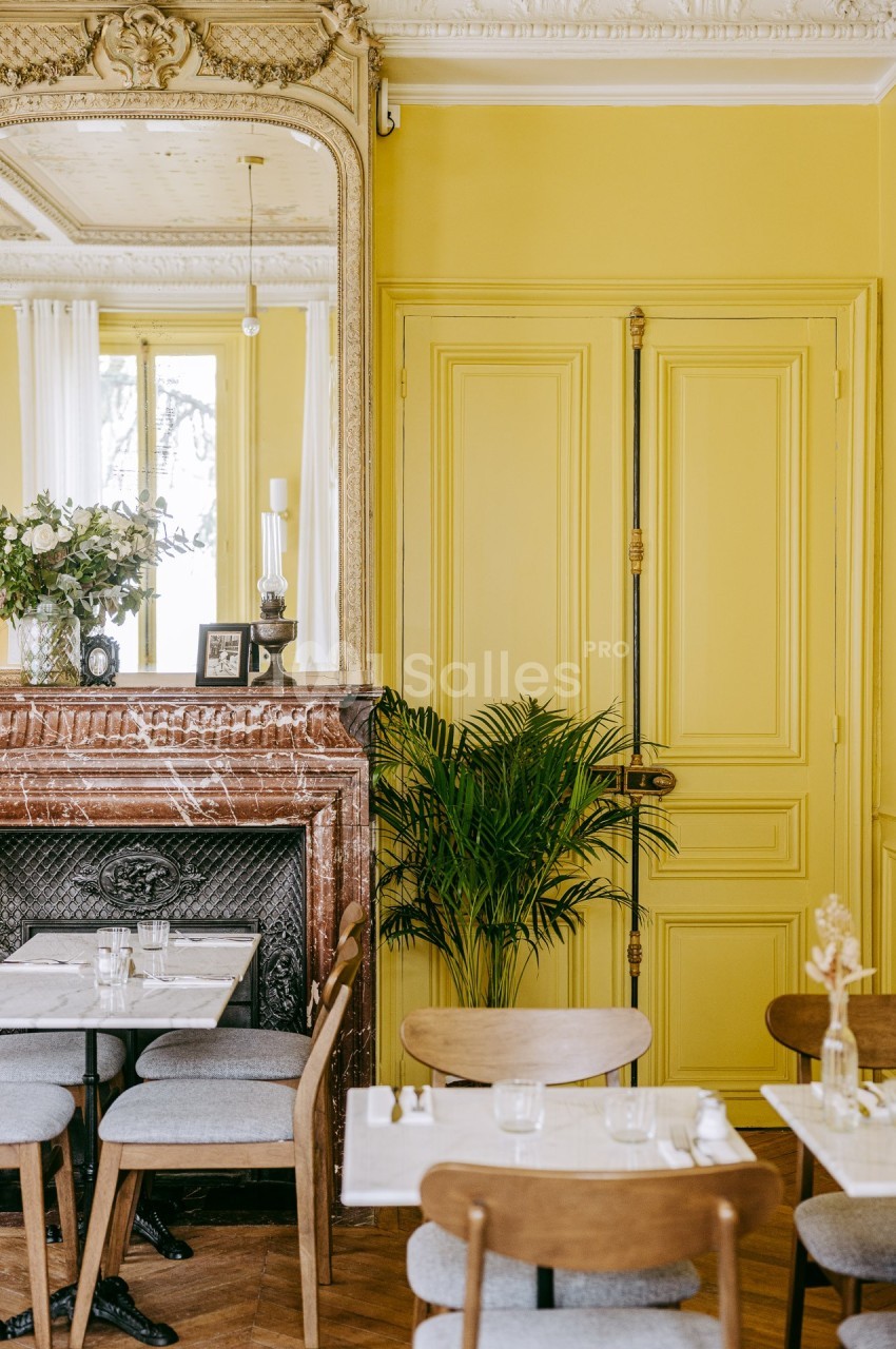 Salle à manger lumineuse avec murs jaunes, cheminée en marbre, miroir orné et table dressée pour deux.
