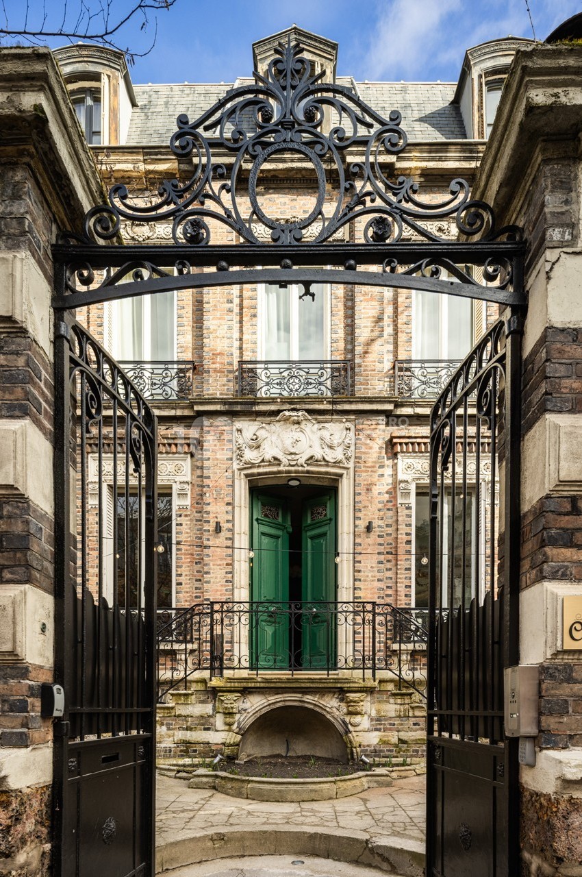 Portail en fer forgé ouvert donnant sur une façade en briques avec balcon et porte d'entrée verte.
