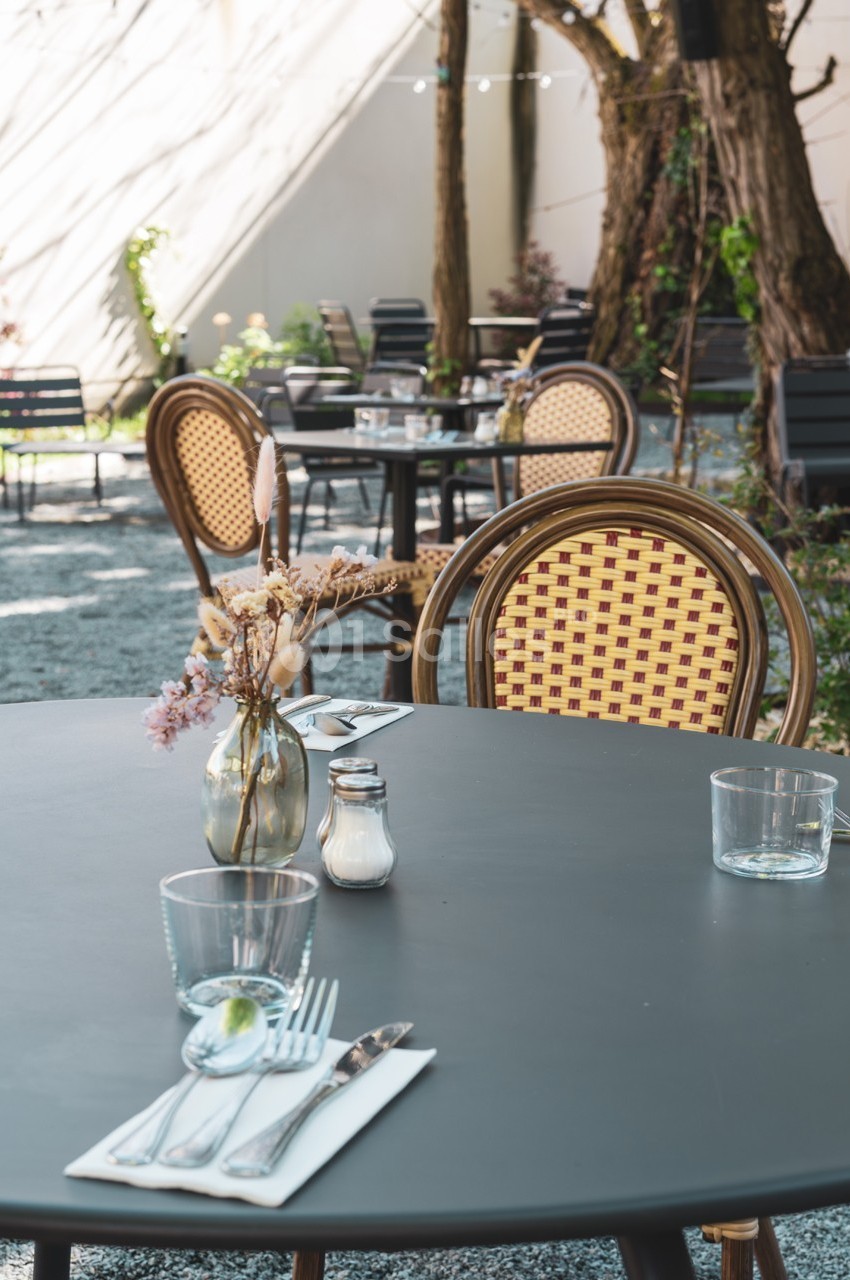 Terrasse extérieure avec tables rondes, chaises en rotin, vase de fleurs séchées et ambiance calme sous des arbres.