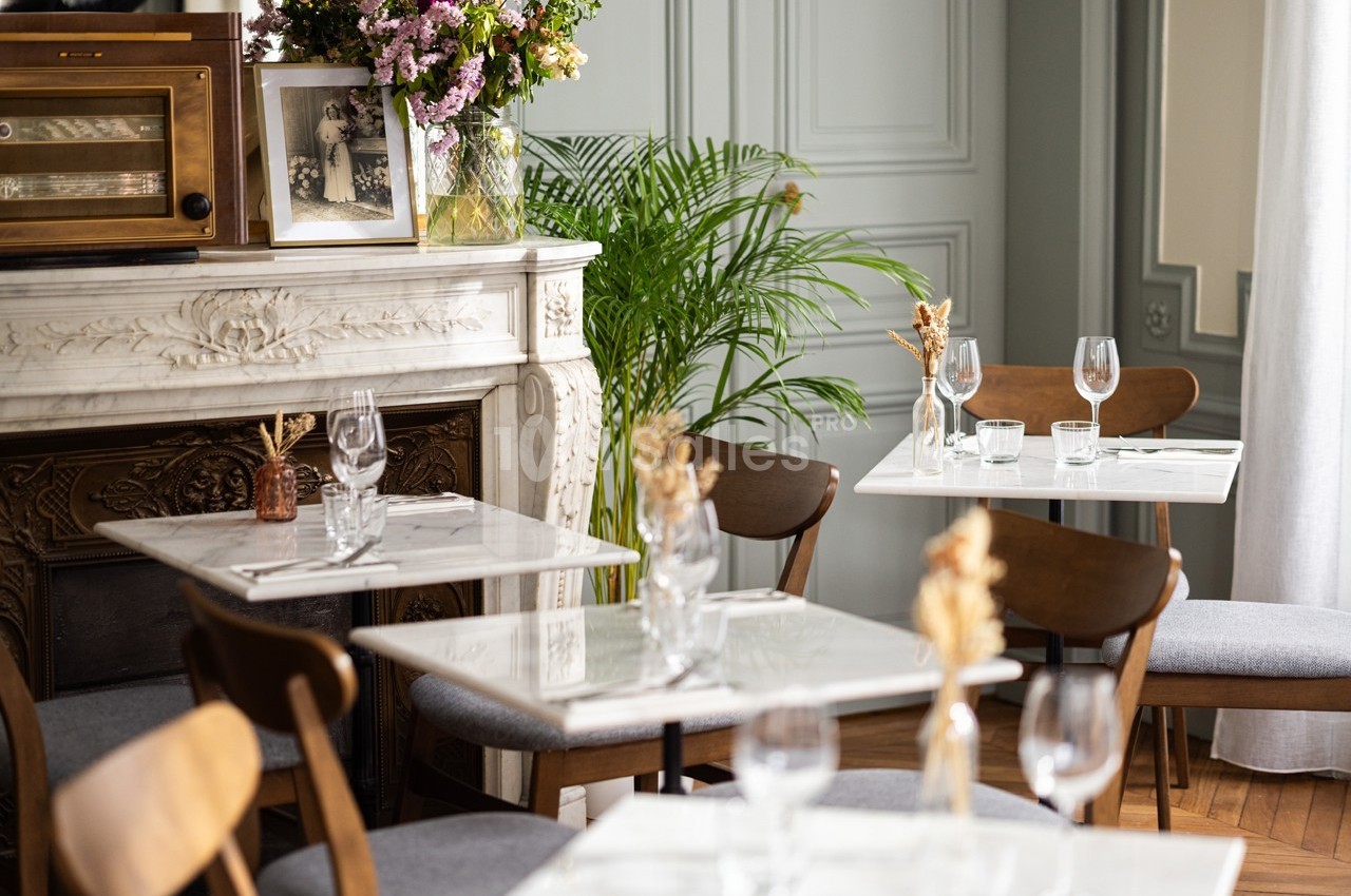 Salle de restaurant lumineuse avec tables en marbre, chaises en bois, décoration florale et cheminée ornée.