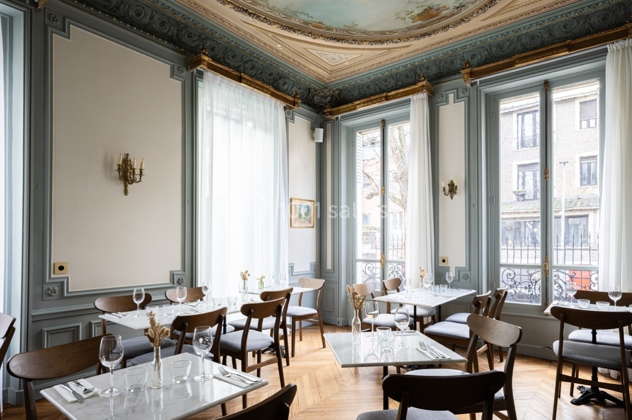 Salle de restaurant élégante avec moulures au plafond, grandes fenêtres, tables dressées et chaises en bois.