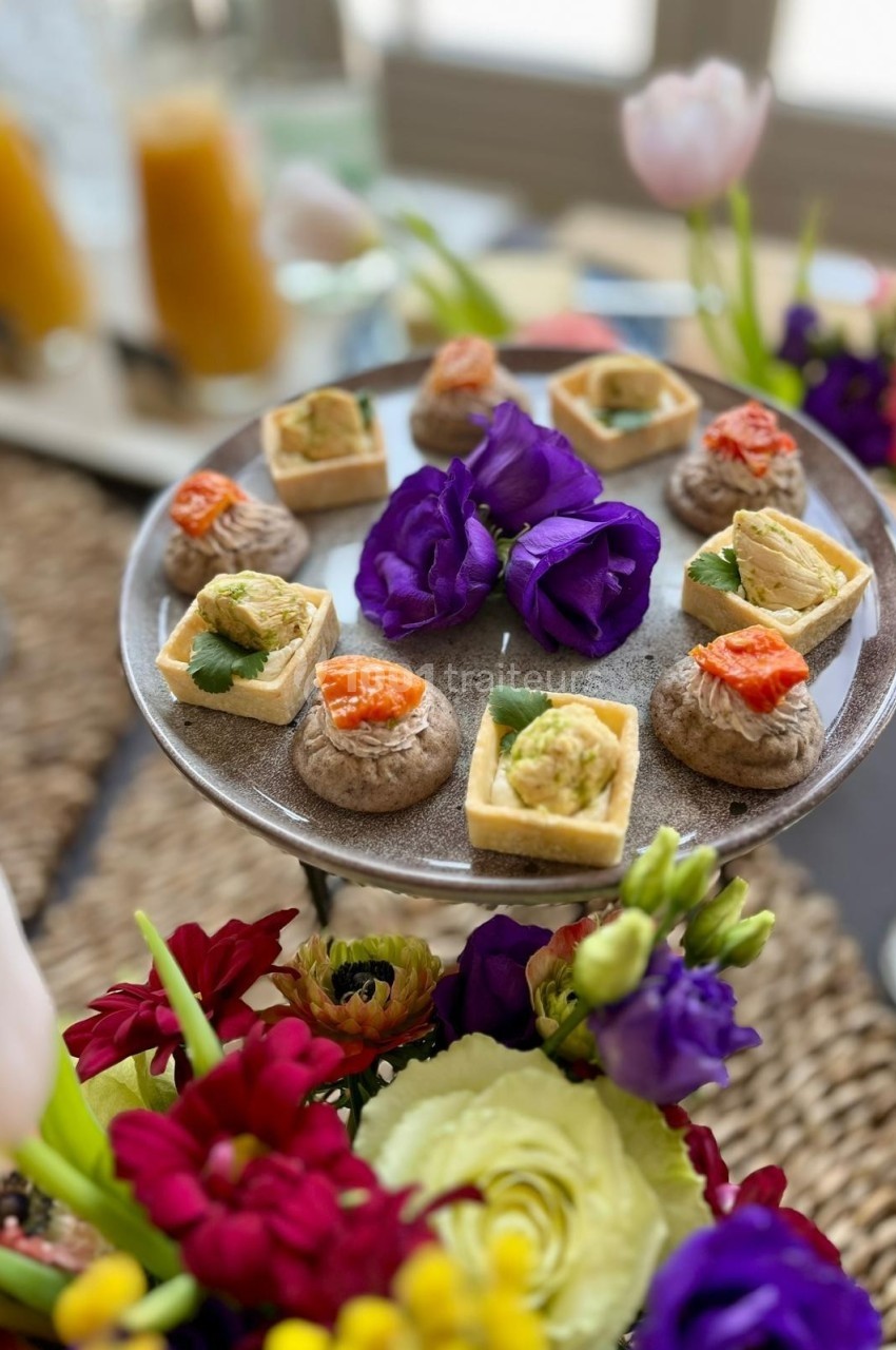 Plateau de bouchées apéritives variées entouré de fleurs colorées, avec des boissons en arrière-plan.
