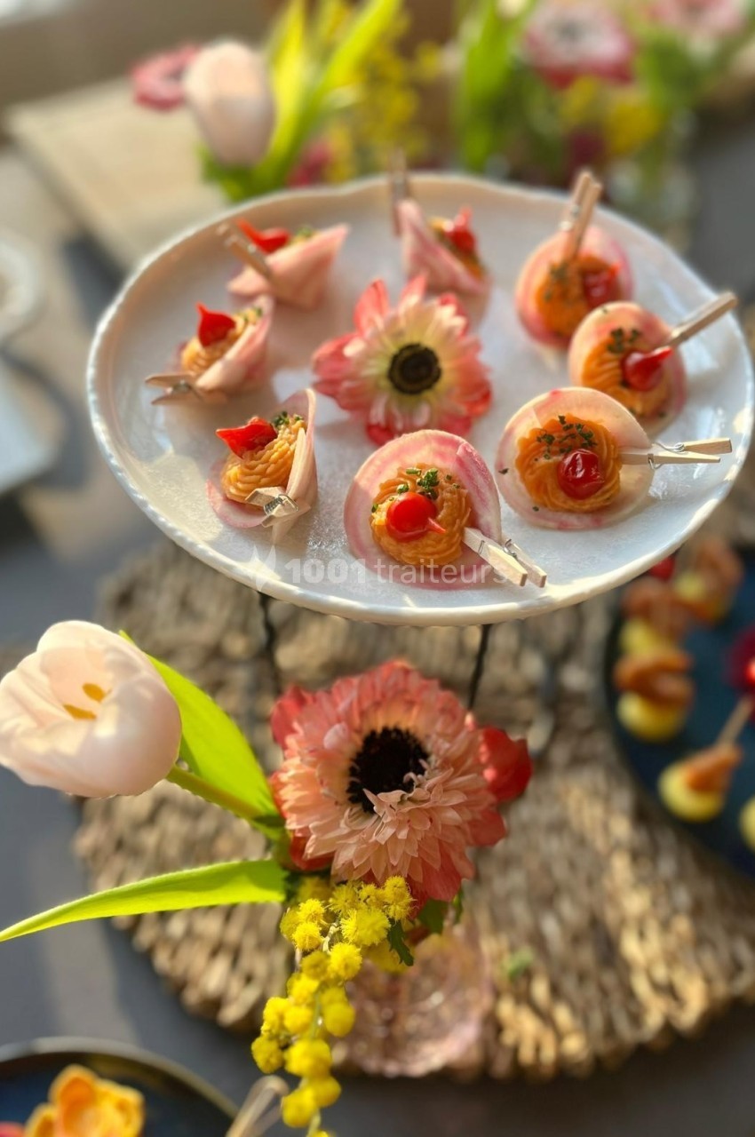 Plateau de bouchées apéritives colorées sur un présentoir, entouré de fleurs décoratives sur une table.