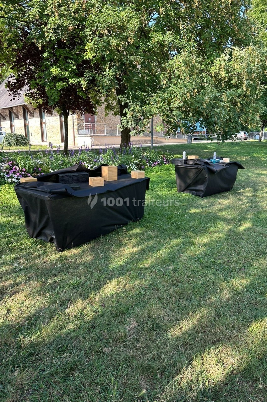 Deux tables recouvertes de nappes noires avec des objets en bois et des accessoires, disposées sur une pelouse ombragée.