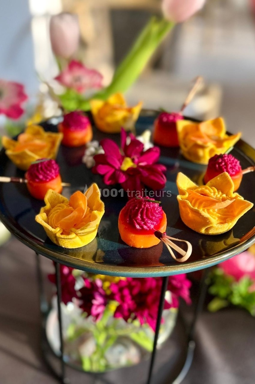 Plateau de bouchées colorées et décoratives, présentées sur un support noir entouré de fleurs fraîches.