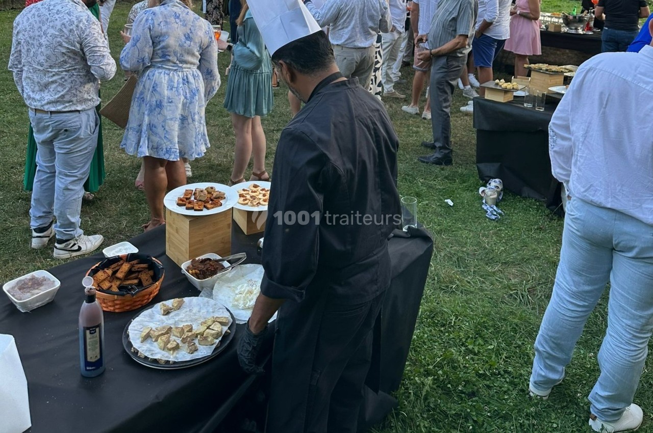 Un chef en tenue noire prépare un buffet en plein air lors d'un événement avec des invités en arrière-plan.