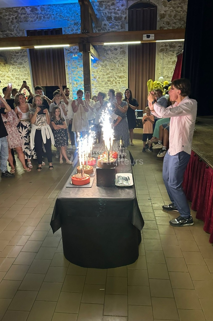 Un homme souffle les bougies sur plusieurs gâteaux avec des cierges magiques, entouré d'un groupe de personnes applaudissant.