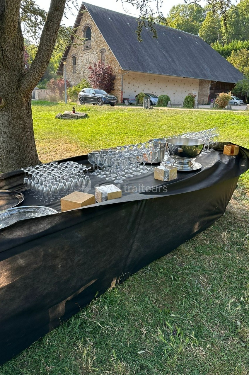Table extérieure dressée avec des verres et des plateaux, située dans un jardin près d'une grange en pierre.