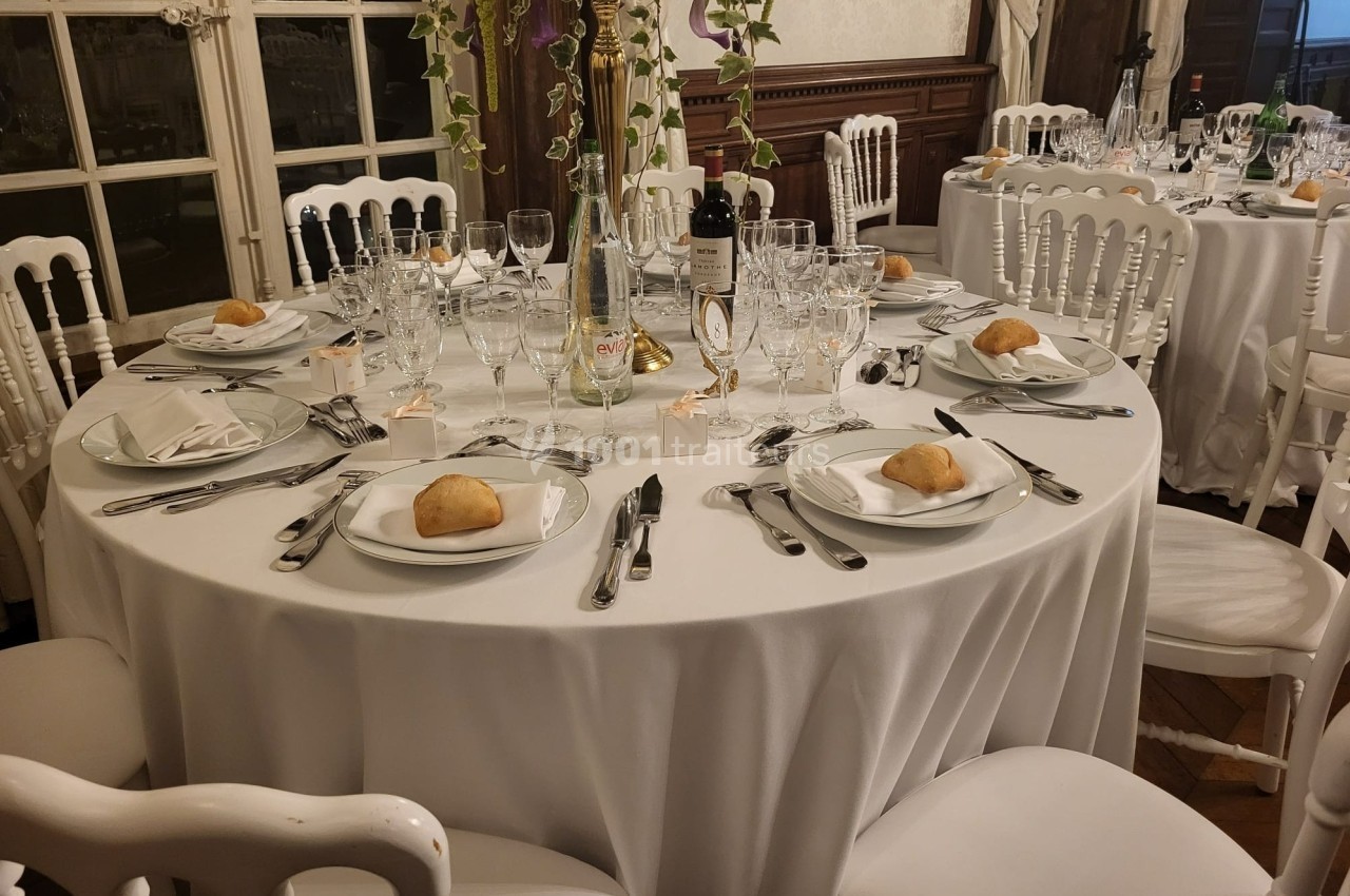 Table ronde élégamment dressée avec nappes blanches, couverts, verres, assiettes et petits pains, dans une salle raffinée.