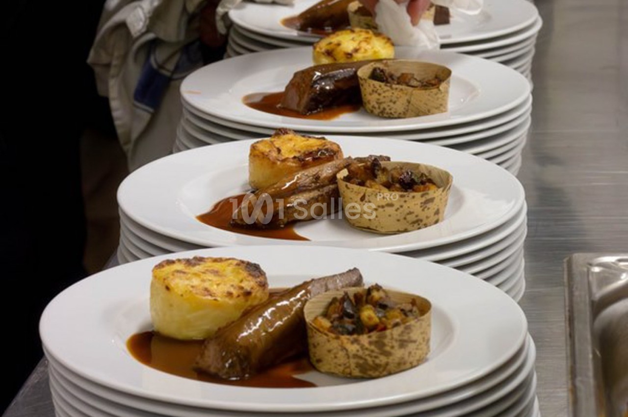Assiettes dressées avec viande, gratin, garnitures et sauce, alignées sur un plan de travail en cuisine.