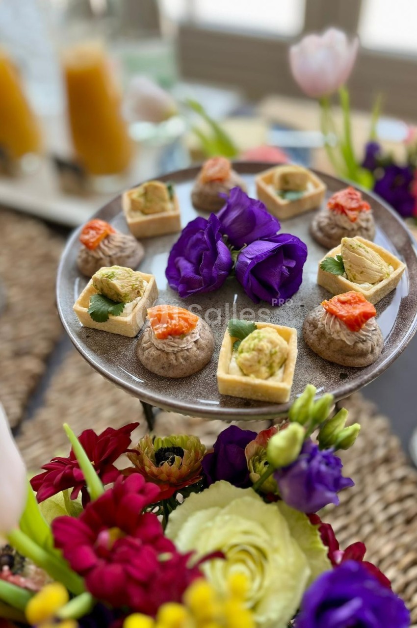 Plateau de bouchées apéritives variées entouré de fleurs colorées, avec des boissons en arrière-plan.
