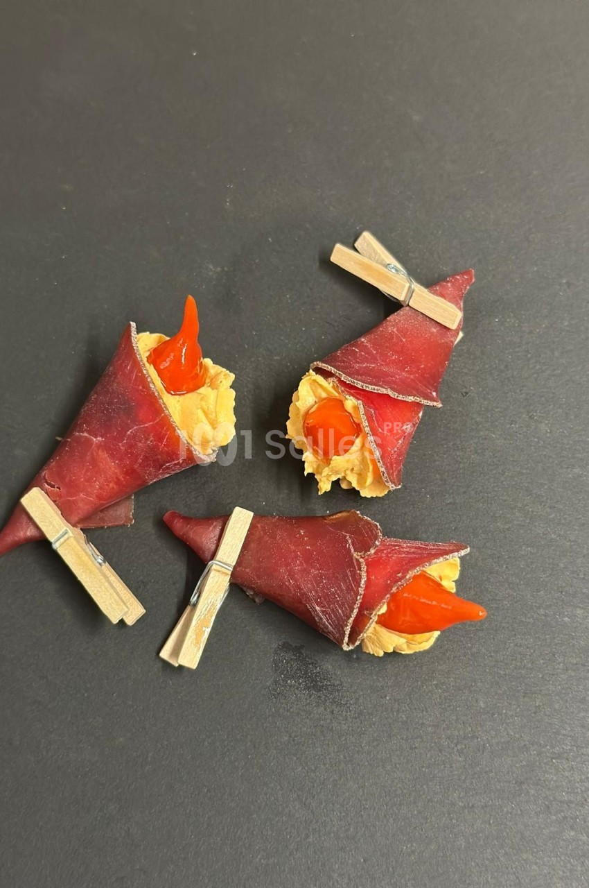 Trois amuse-bouches enroulés dans de la viande séchée, garnis de fromage et décorés de piments rouges, fixés par des pinces.