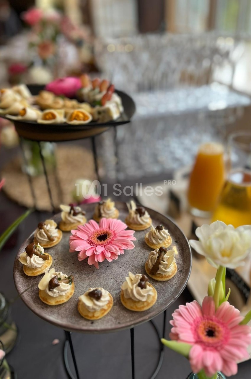 Plateau de petits amuse-bouches décorés, entouré de fleurs et de boissons sur une table élégamment dressée.