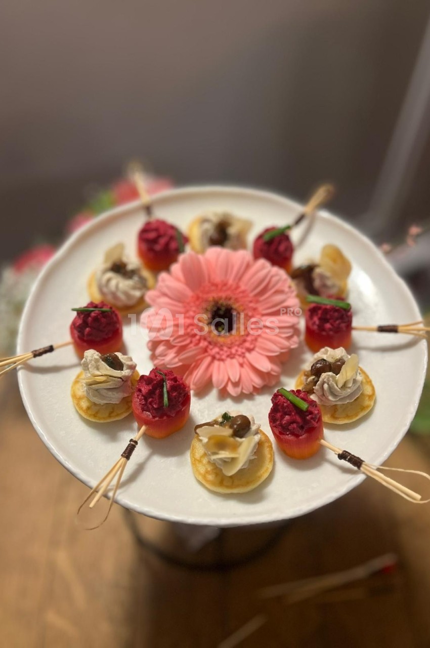 Assortiment de bouchées apéritives colorées disposées autour d'une fleur rose sur un plateau blanc.