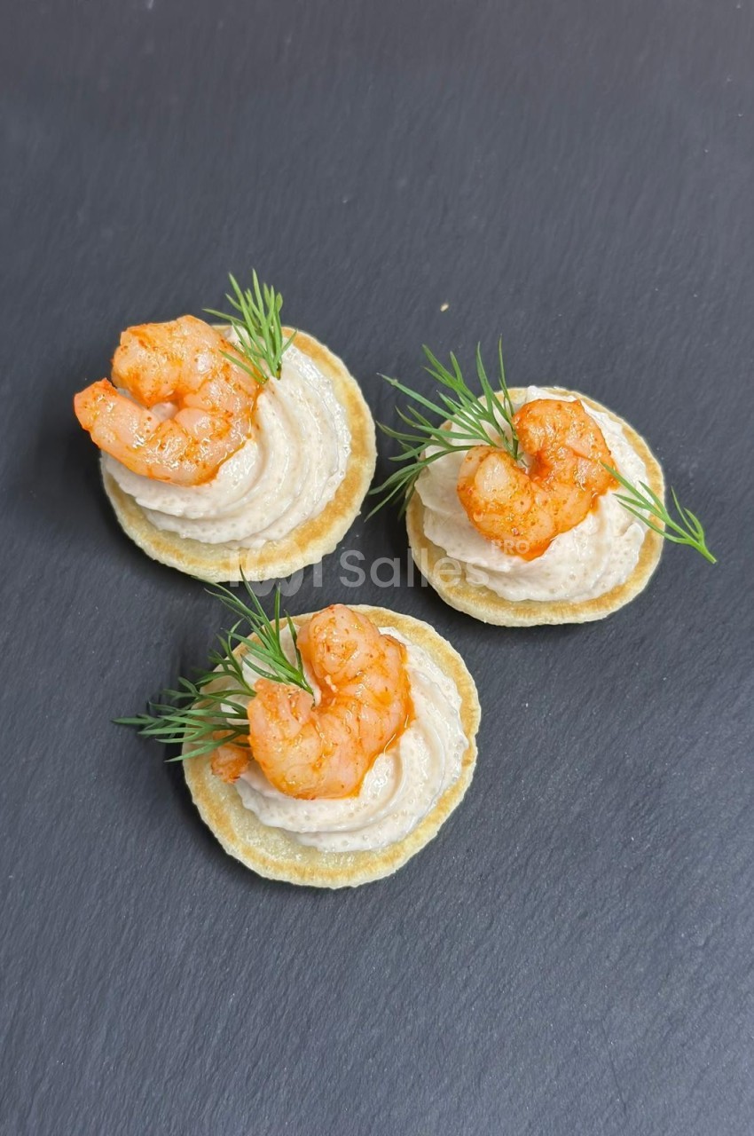 Canapés sur crackers garnis de crème, crevettes épicées et brins d'aneth, présentés sur une ardoise noire.