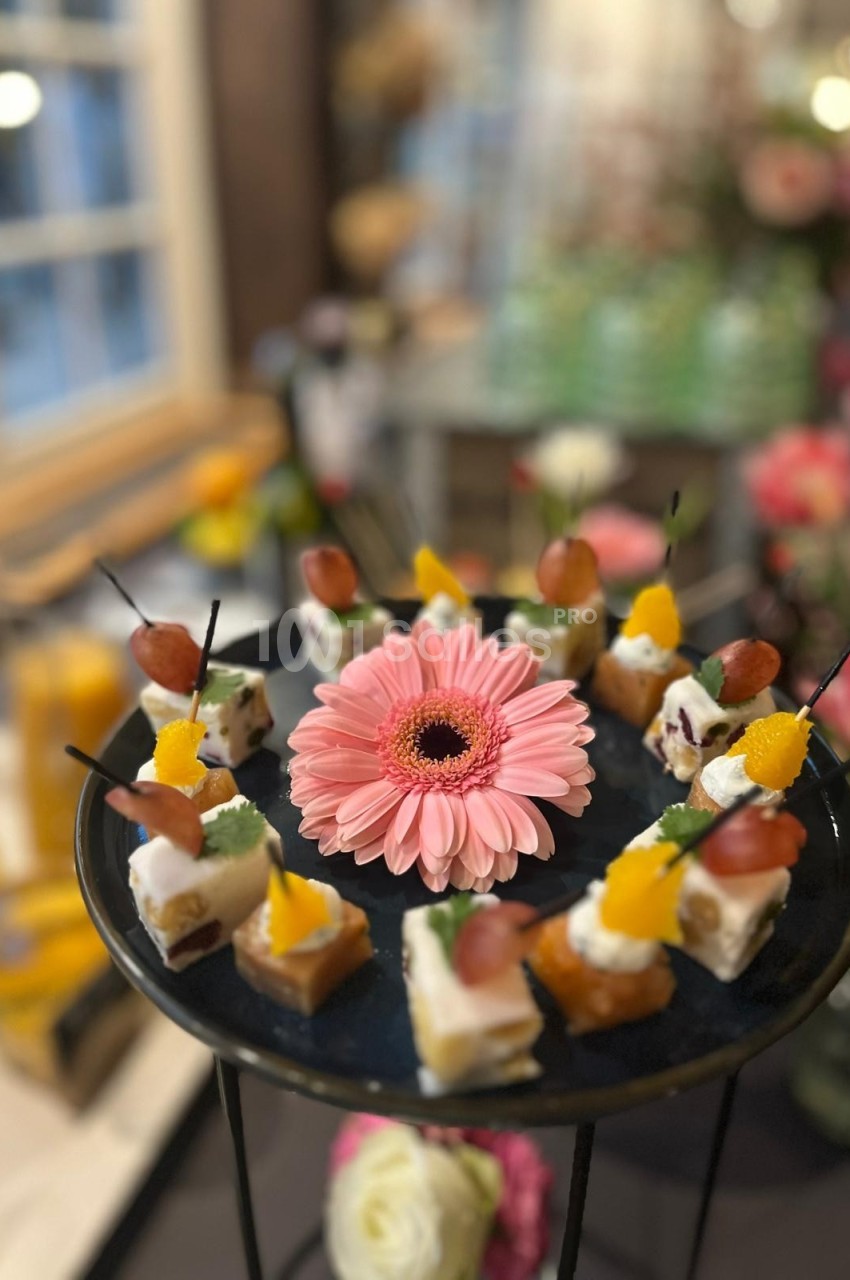 Plateau de bouchées apéritives décoré d'une fleur rose au centre, entouré de verrines en arrière-plan.