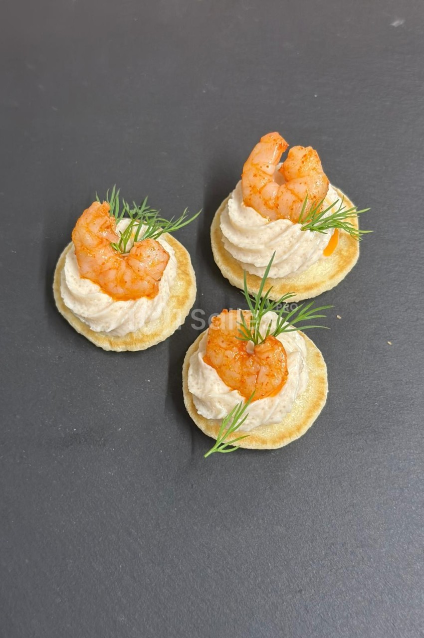 Canapés surmontés de crevettes grillées, mousse crémeuse et brins d'aneth, disposés sur une surface sombre.
