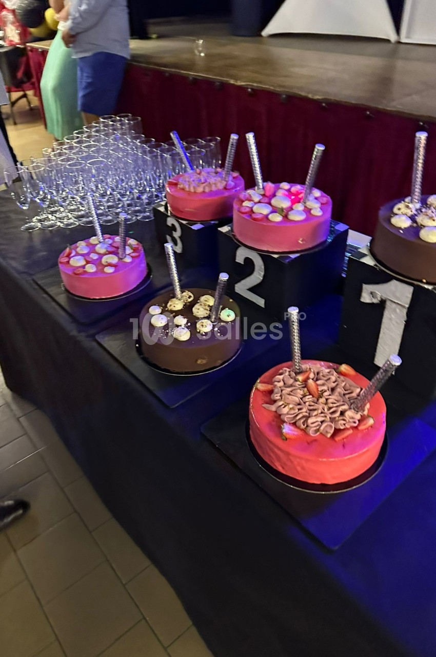 Gâteaux décorés numérotés de 1 à 3 sur une table noire, entourés de verres empilés et d'invités en arrière-plan.