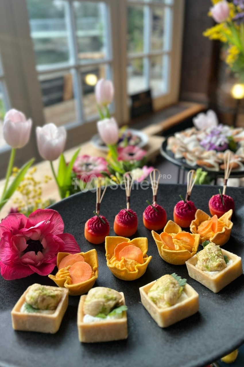 Plateau de bouchées apéritives variées présentées sur une table décorée de fleurs près d'une fenêtre.
