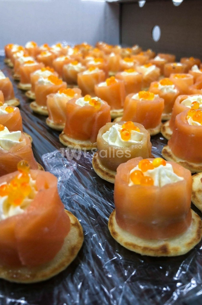 Bouchées apéritives composées de saumon fumé, crème et œufs de poisson sur des blinis disposés en rangées.