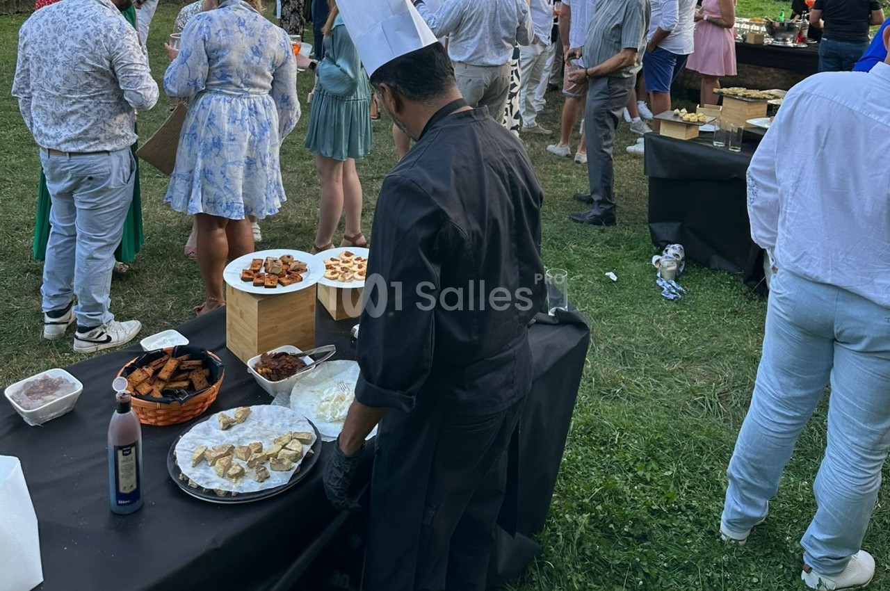Un chef en tenue noire prépare un buffet en plein air lors d'un événement avec des invités en arrière-plan.