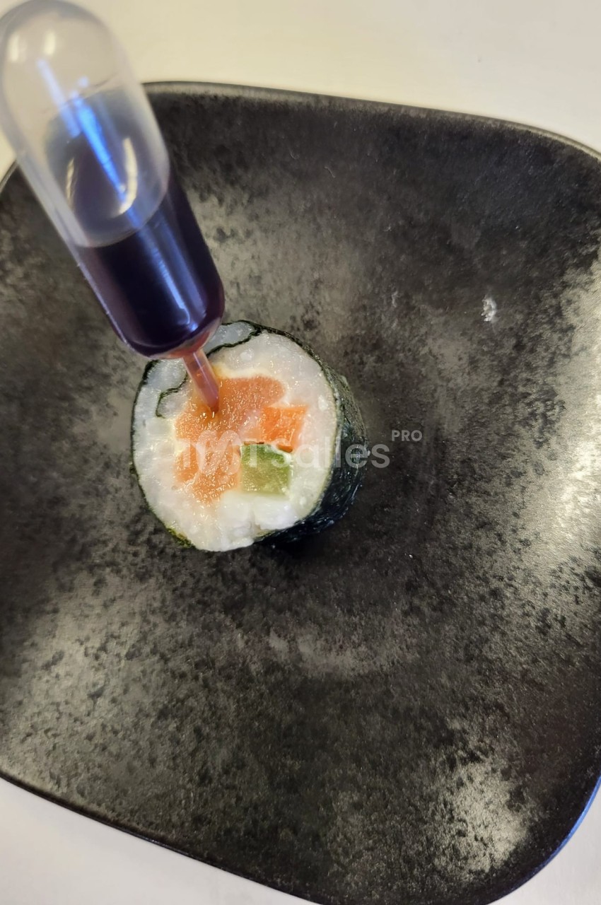 Sushi roulé avec saumon, avocat et riz, accompagné d'une pipette contenant une sauce foncée, présenté sur une assiette noire.
