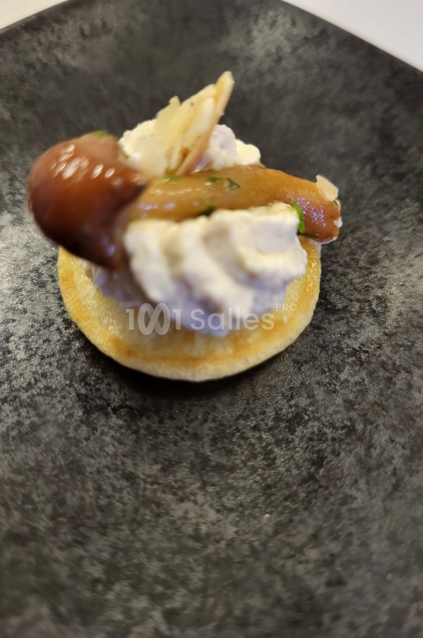 Blinis garni de fromage frais, champignons marinés et copeaux de fromage sur une assiette noire.