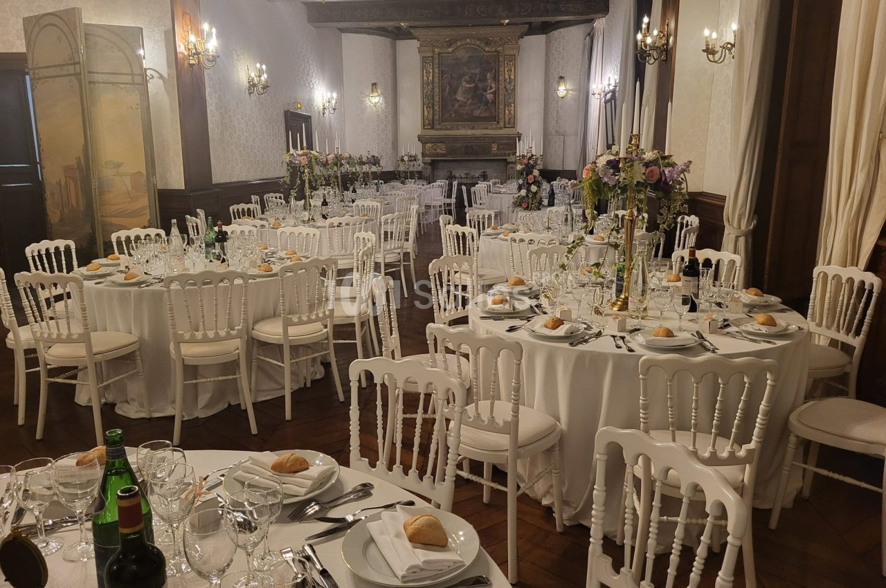 Salle de réception élégante avec tables rondes dressées, décor floral et chaises blanches.