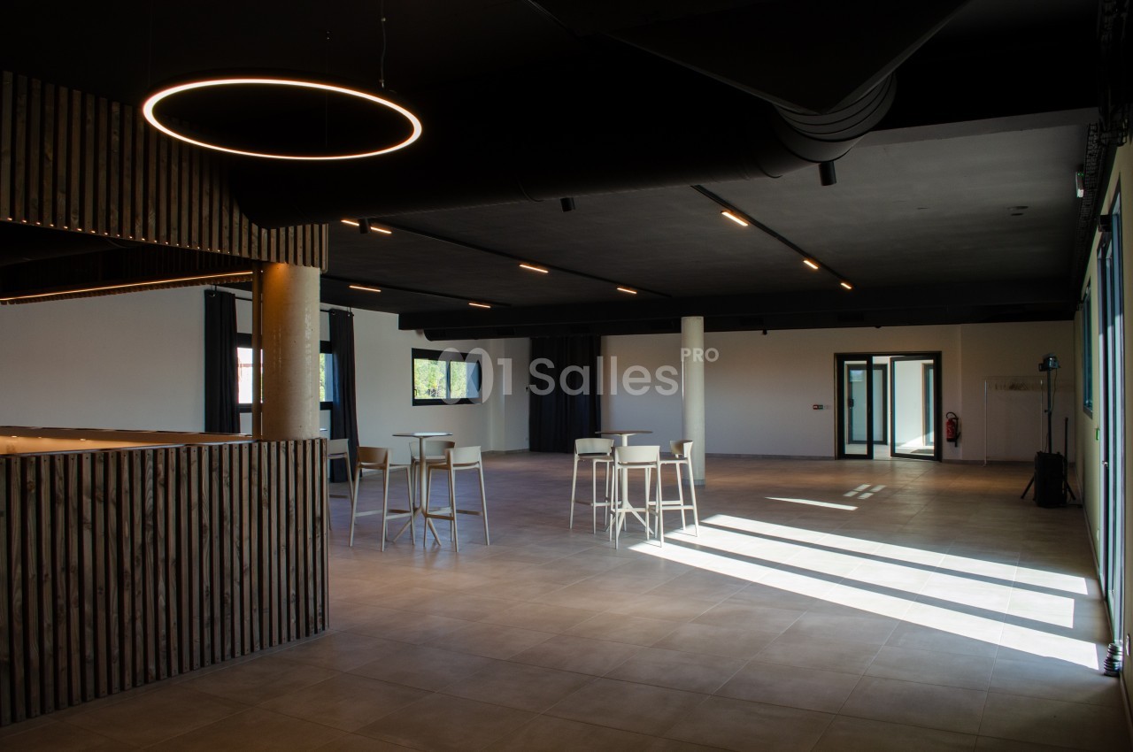 Salle spacieuse avec un éclairage moderne, des tables hautes et un sol carrelé, baignée de lumière naturelle.