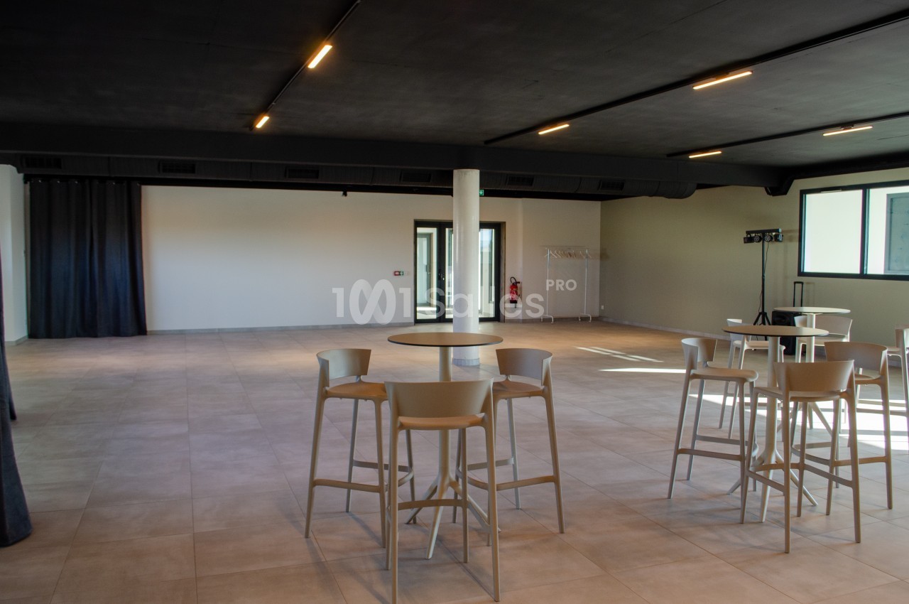 Salle spacieuse et lumineuse avec tables hautes, chaises et sol en carrelage, éclairée par des lumières au plafond.