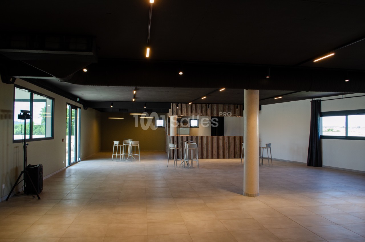 Salle vide avec sol en carrelage, murs clairs, plafond sombre, grandes fenêtres et quelques tables hautes avec tabourets.