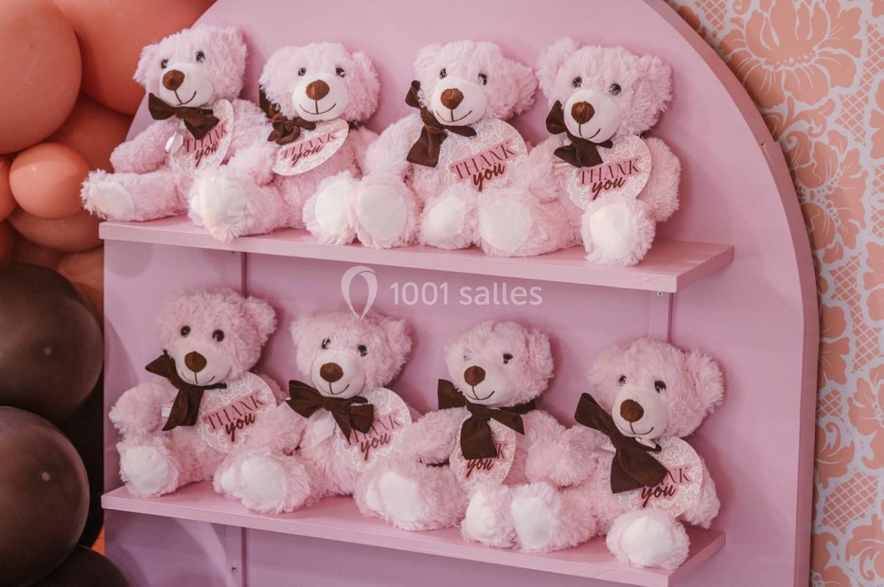 Peluches roses en forme d'ours alignées sur des étagères roses, avec des nœuds marron et l'inscription ’Thank you’.