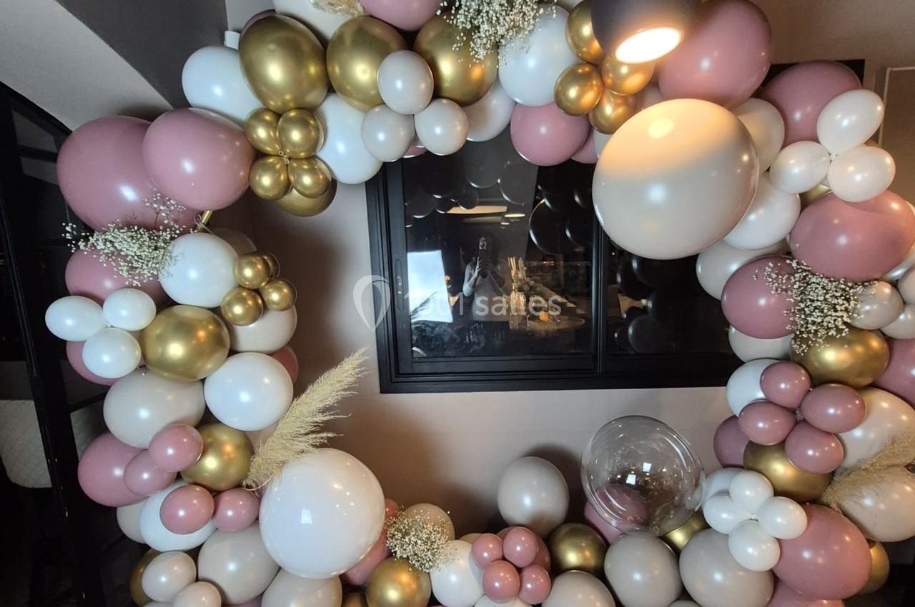 Arche décorative composée de ballons roses, blancs et dorés, ornée de fleurs séchées, dans un intérieur.