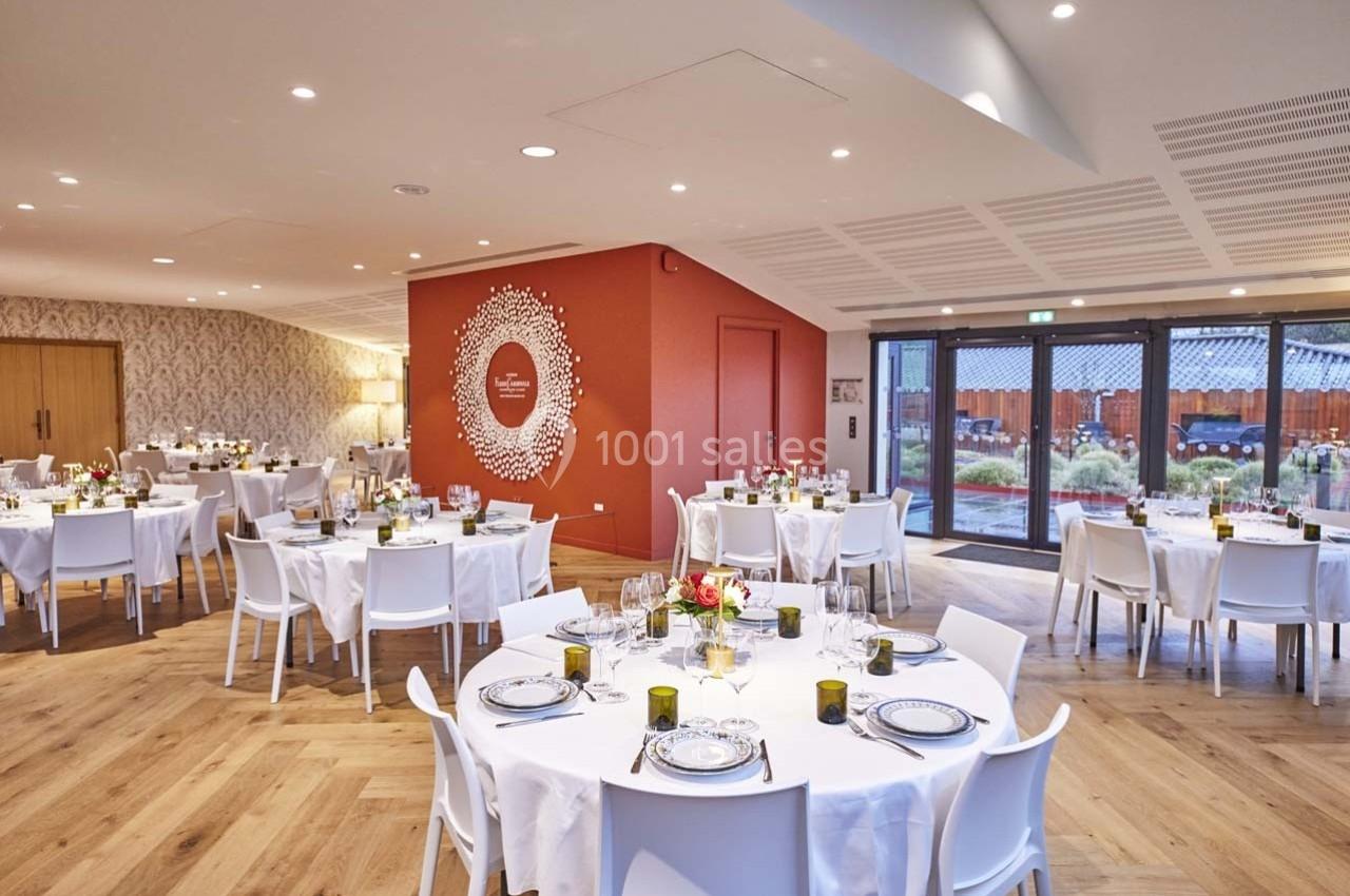 Salle de réception lumineuse avec tables rondes dressées, chaises blanches et décoration murale rouge.