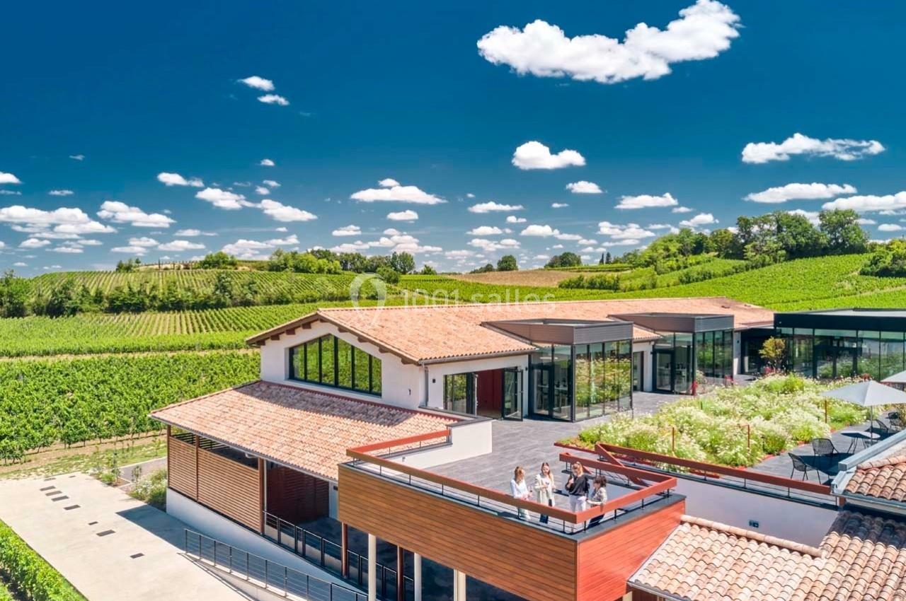 Bâtiment moderne entouré de vignes sous un ciel bleu avec terrasse et grandes baies vitrées.