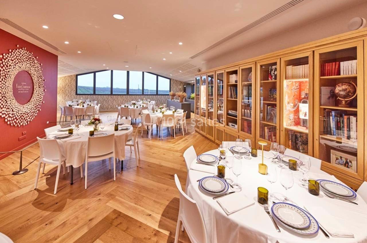 Salle lumineuse avec tables dressées pour un repas, parquet en bois, étagères remplies de livres et grande baie vitrée.